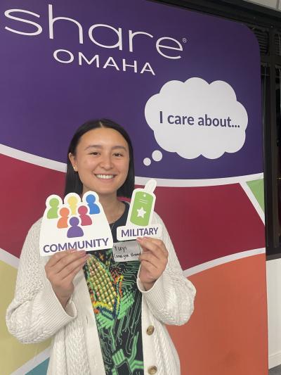 Woman in front of a backdrop that says "I care about" holding signs that say "community" and "military"